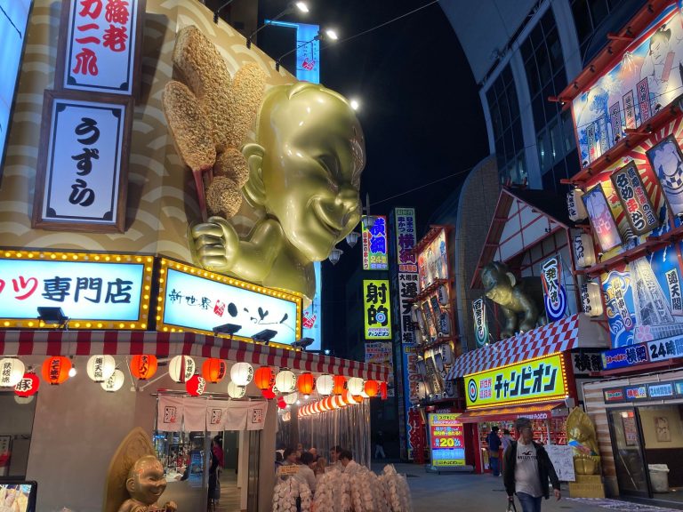 Quartier rétro de Shinsekai à Osaka, avec en premier plan un restaurant de brochettes frites doré très imposant avec des lampions rouges et blancs, et en arrière plan des restaurant aux énormes devantures très colorées, typiques d’Osaka